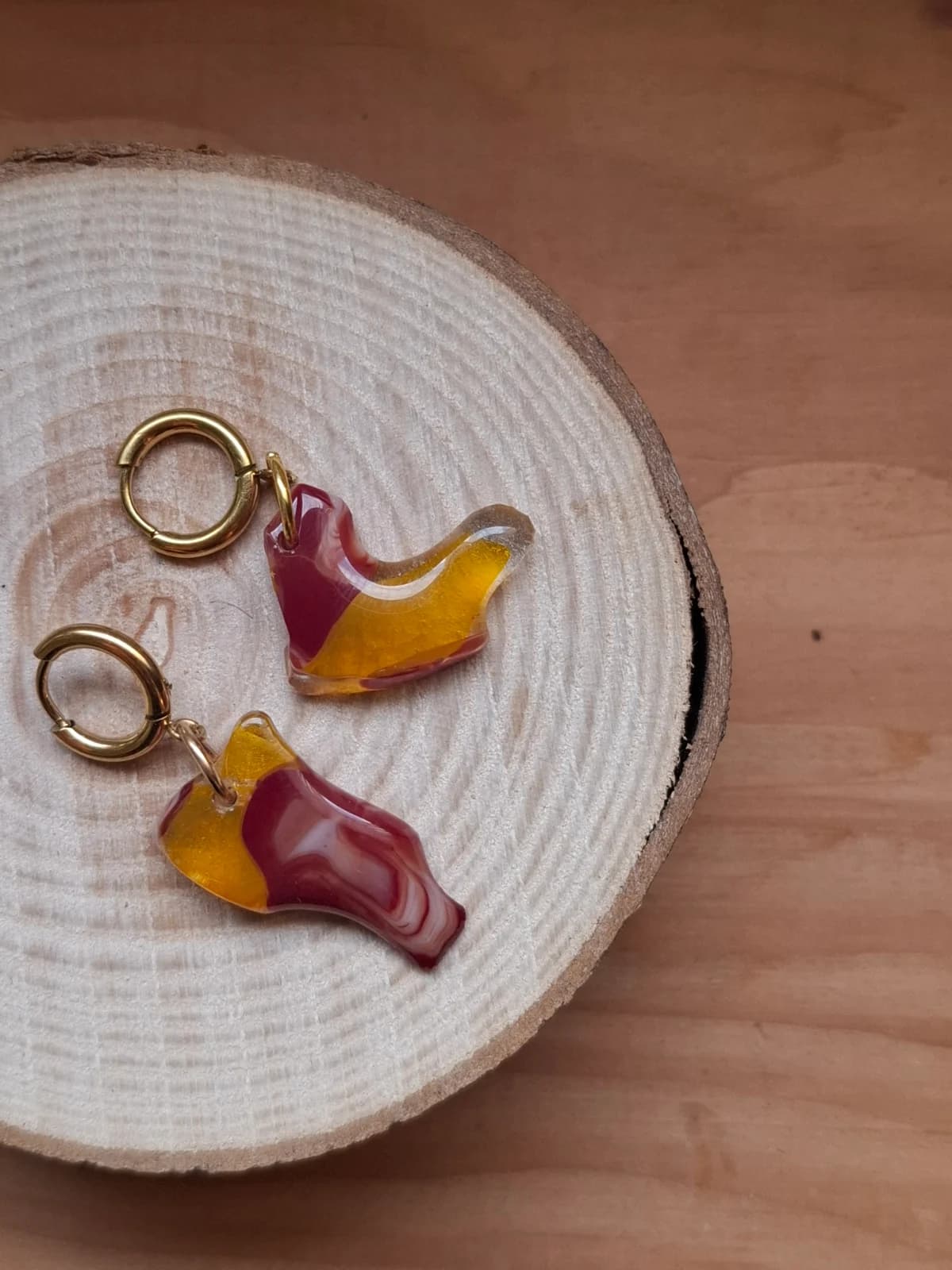 Boucles d'oreilles pendantes asymétriques en verre jaune et rouge posées sur une rondelle de bois.