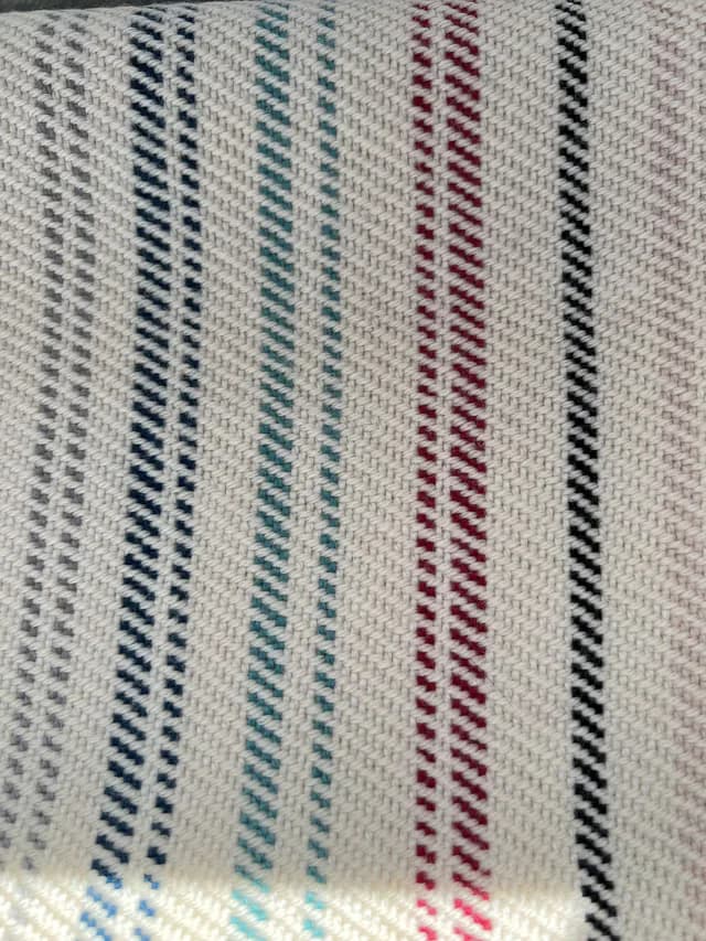 Gros plan sur le tissage de l'écharpe avec rayures fines verticales bleues, bordeaux et noires sur fond écru.