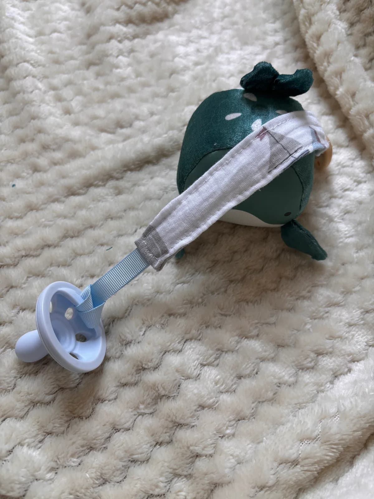 Vue de dessus de l'attache-tétine en tissu avec passant bleu relié à une sucette blanche sur fond doux.