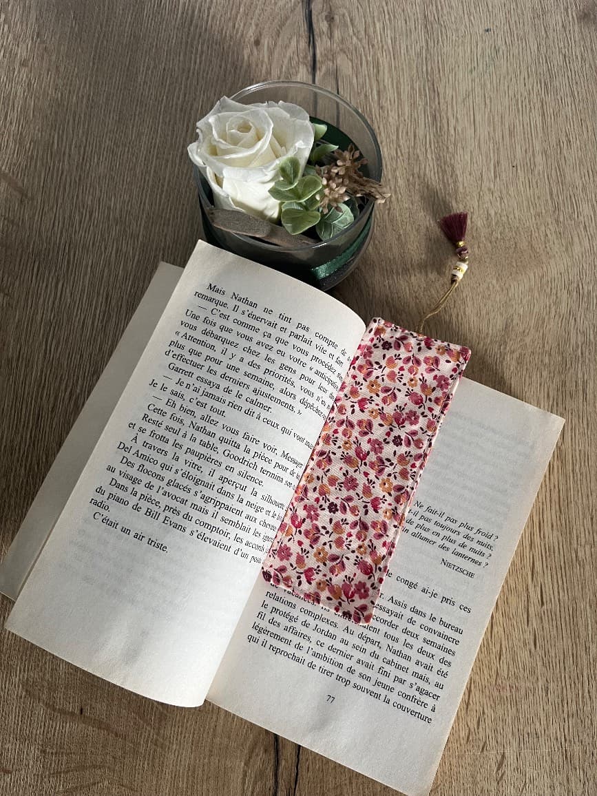 Marque-page en tissu fleuri rose et son pompon posés entre les pages d'un livre ouvert.