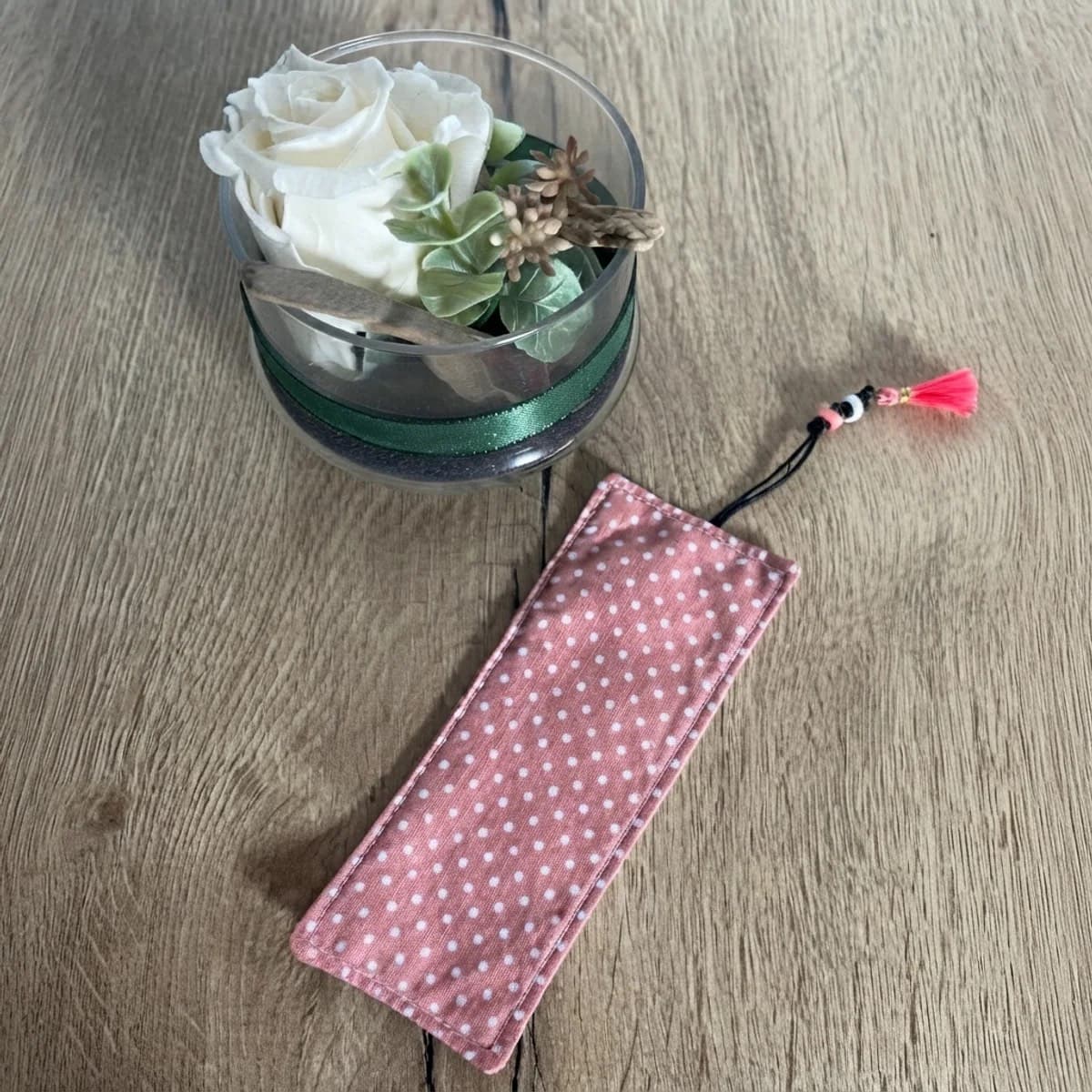 Marque-page plat en tissu rose à pois blancs avec cordon noir et pompon rose, posé sur une table en bois.