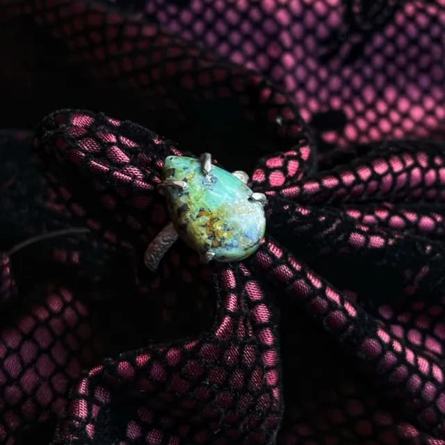 Bague réglable en argent avec pierre jaspe en goutte verte et brune posée sur un tissu noir et rose.