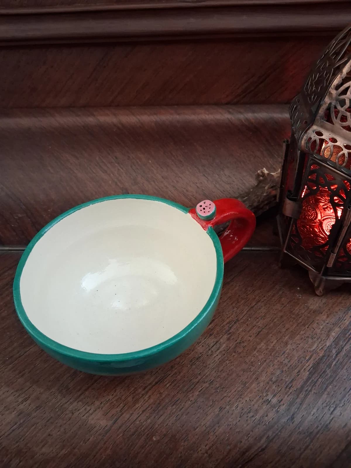 Tasse émaillée verte à intérieur blanc et anse rouge ornée d'un détail pastèque, posée sur du bois.