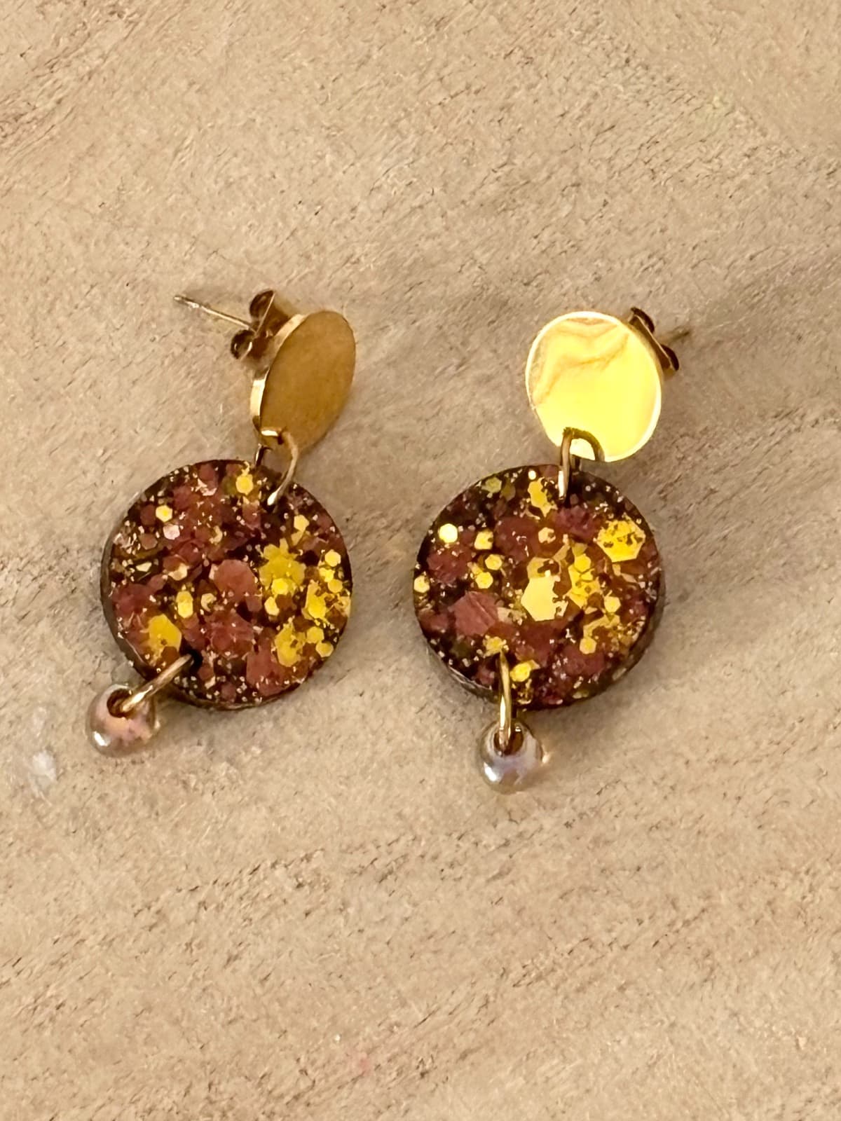 Boucles d'oreilles dorées avec médaillon rond pailleté marron posées à plat sur du bois